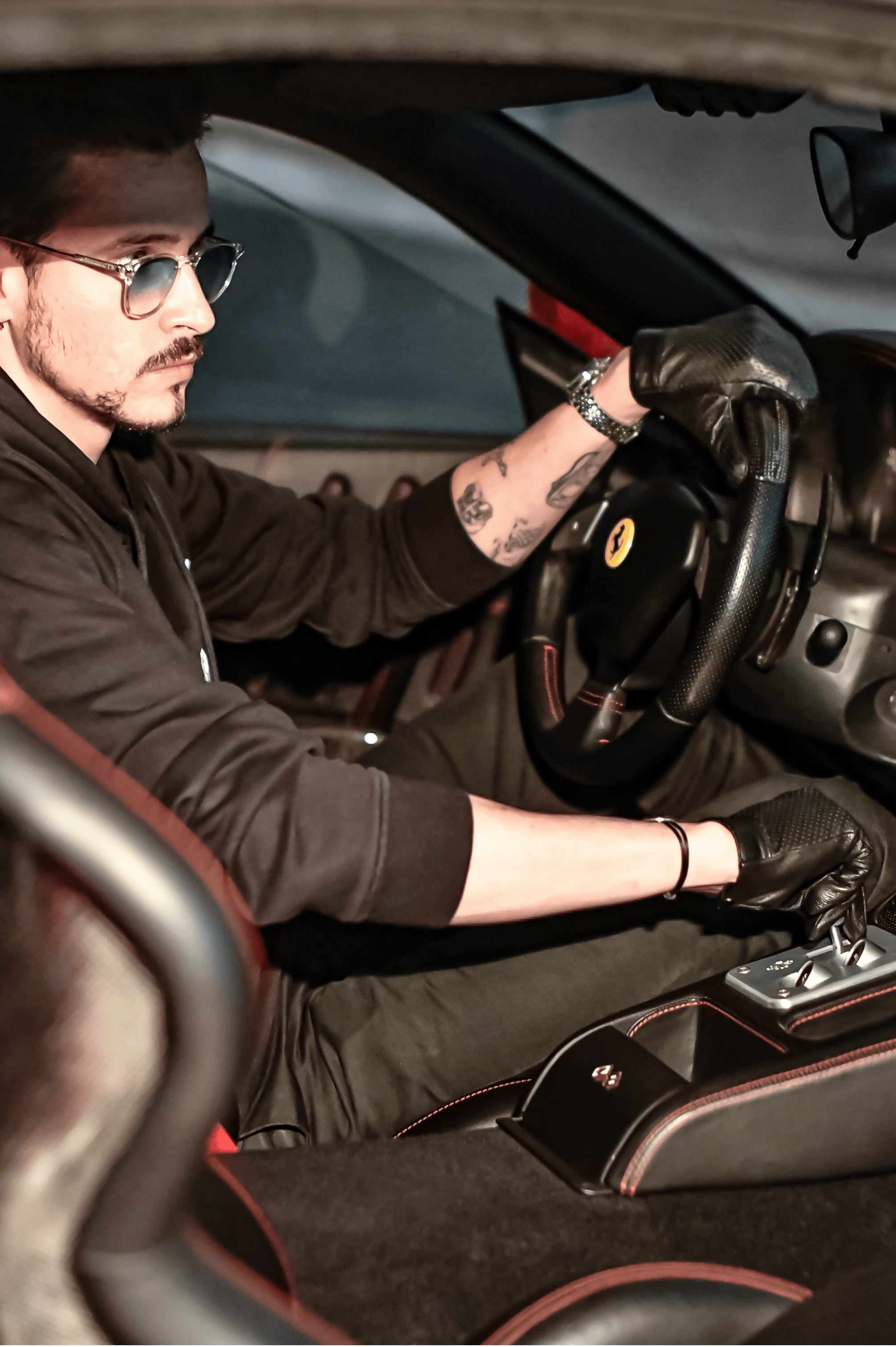 yann delplanque portant des gants de conduite noir profond pour homme au volant d'une ferrari intérieur noir surpiqures rouges - La Main De l'Homme